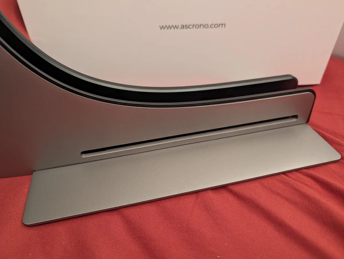 The empty ASCRONO dock in side profile on a red cloth, showing the machined slot the MacBook Pro drops into and the wide flat base plate, with the product box behind it