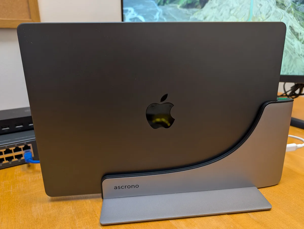 The rear of the docked MacBook Pro, with the ascrono wordmark on the base and a small green status light glowing at the top of the connector block