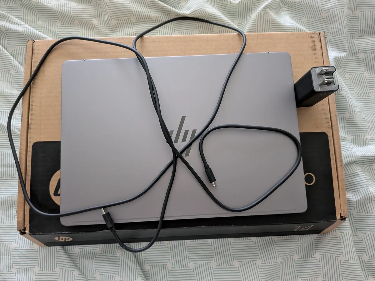 HP Omnibook X laptop on its box with USB-C cable and power adapter on a patterned bedspread.