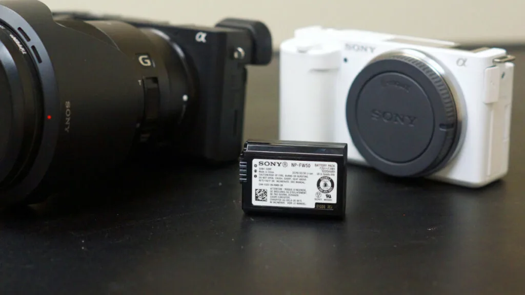 Sony NP-FW50 battery pack in foreground, with Sony a6500 and ZV-E10 cameras blurred in background.